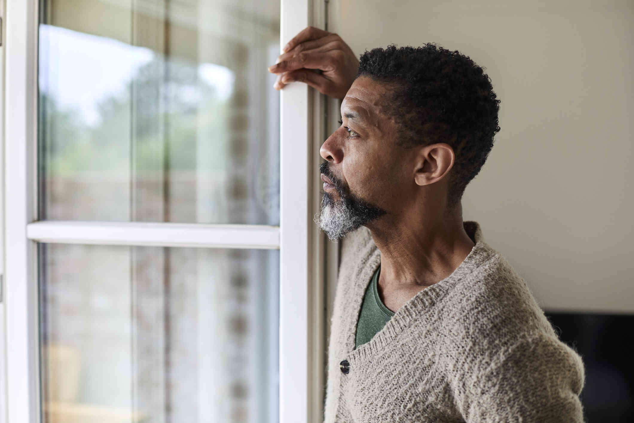 A close up of a middle aged man as he leans against a window frame and gazes out of the window while deep in thought.